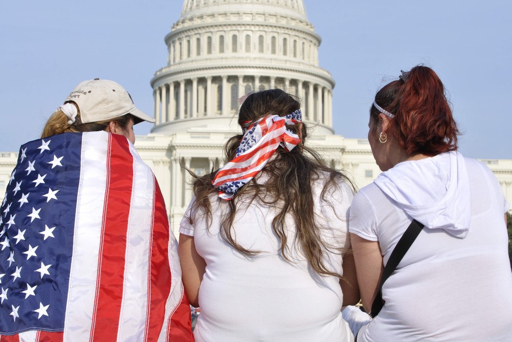The backs of three people who are facing the U.S. Capitol Building, two of them wear American flags
