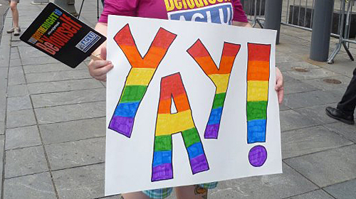 A person holding a sign reading "Yay!" in rainbow letters.
