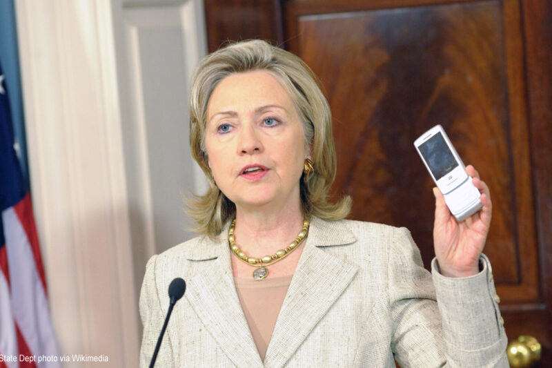 Hillary Clinton as Secretary of State at podium holding a cell phone