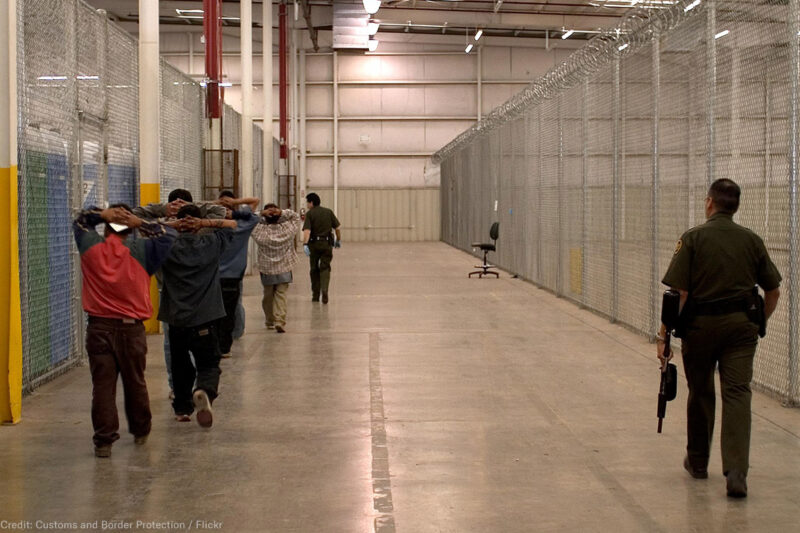 Immigrant detainees walk down a hall escorted by heavily armed CBP officers.