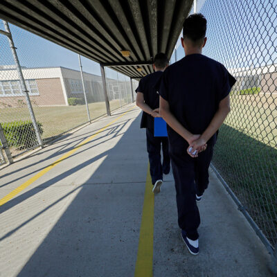 Immigrant detainees walk with their hands clasped behind their backs along a line painted on a walkway