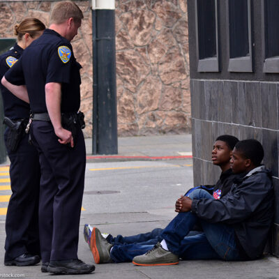 Two San Francisco police officers interrogating and standing over two Black American men seated on the sidewalk. Overall, Black Americans are arrested at 2.6 times the per-capita rate of all other Americans.