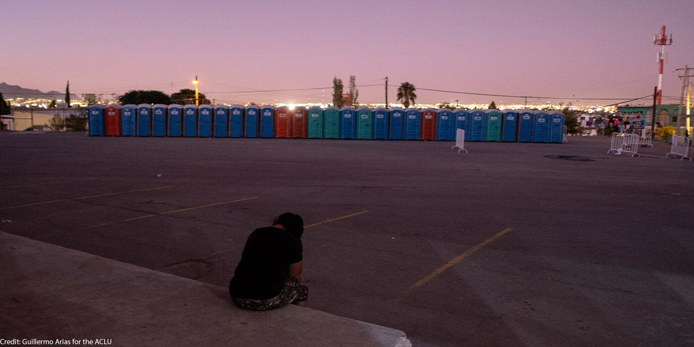 A woman sits outside in an empty lot in Ciudad Juarez, Mexico at sunset, with her head hanging down, and a line of outhouses across the lot.