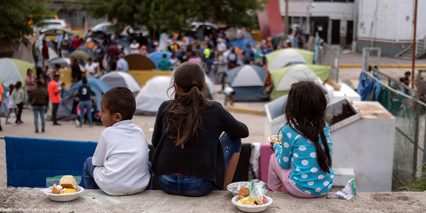 Three children sit down while eating food, backs turned to the camera. In front of them stand men, women, and children in a lot full of tents where they stay. Taken in Matamoros, Mexico.