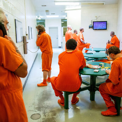 Inmates pass the time in their cell block. Some pictured on the phone, some watching television, and some playing card games at tables. As concerns regarding the spread of COVID-19 mount, inmates argue that prisons have no plan in place protect against the spread of the illness.