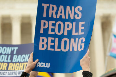 Someone holding up a poster in front of the Supreme Court that reads "trans people belong" and in the background, another person holds a poster that says "don't roll back our rights." This is in the face of trans discrimination and at a time when SCOTUS has a number of cases on the docket that affect trans lives.