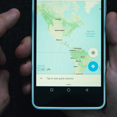 One hand holding a cell phone while the other hovers with index finger extended over a smart phone showing a map of North and South America.