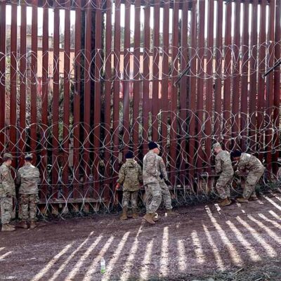 US military members are seen next to a red metal and barbed wire fence at the US border