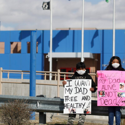 Two children wear masks and hold signs that read "I want my dad free and healthy" and "I want my dad alive not dead!!" as part of a protest calling for the release of detained immigrants in front of the GEO Detention Center in Aurora, Colorado.