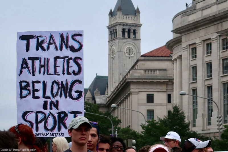 A crowd of marchers with one holding a sign with the text " Trans Athletes Belong in Sport."