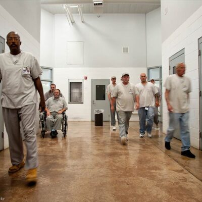 Prisoners are seen walking in a hallway in a prison, one in a wheelchair