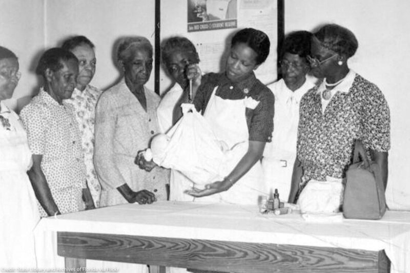 A black nurse showing black midwives how to use a portable scale to weigh babies.