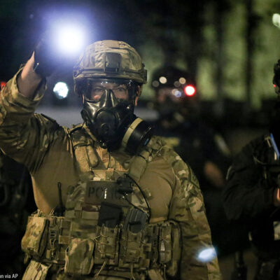 A group of militarized federal agents with an agent in the center shining a bright flashlight toward the viewer.