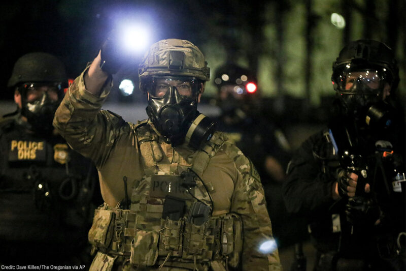 A group of militarized federal agents with an agent in the center shining a bright flashlight toward the viewer.