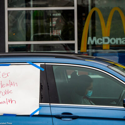 A protester in a car with a sign reading "Worker Health is Public Health" at a socially distanced protest.