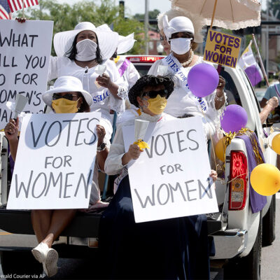 Demonstrators in period clothing with signs advocating for women's suffrage.