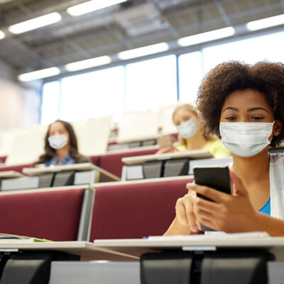 A girl sits in a college lecture hall, wearing a mask and scrolling through phone in hand.