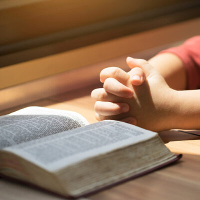 A boy whose hands are folded in prayer in front of a Bible on a desk.