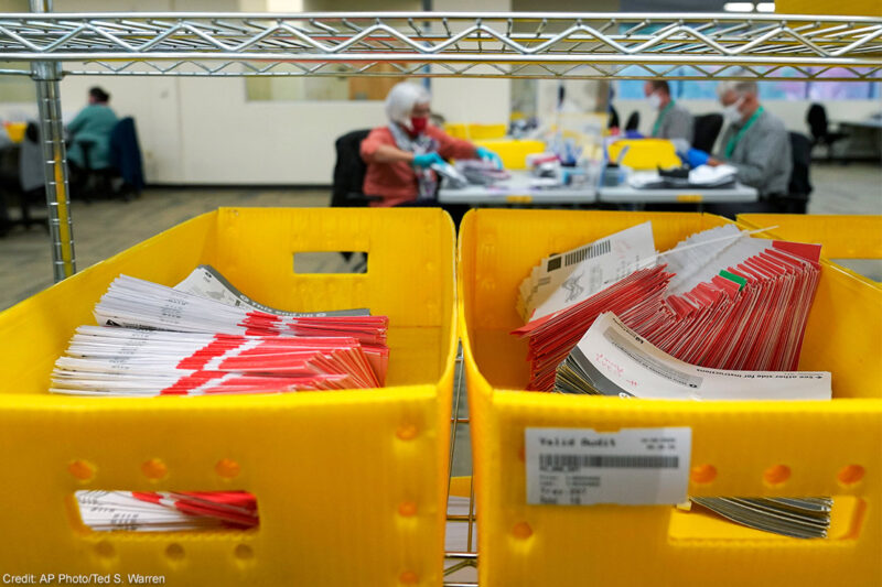 Boxes of vote-by-mail ballot envelopes with workers counting ballots in the background.