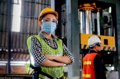 A technician wearing a surgical mask and hard hat stands with arms crossed in a factory.