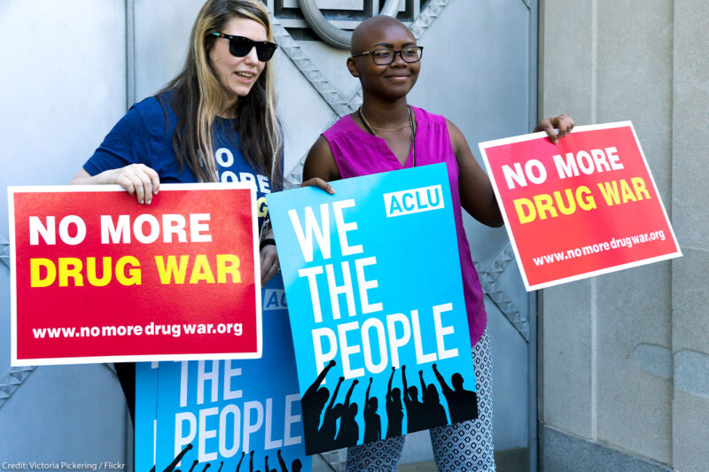 Two protestors holding 'No More Drug War' and ACLU's 'We the People' posters at a protest in front of the Justice Department.