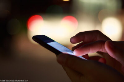 A pair of hands holding a cell phone at night with street lights in the background.