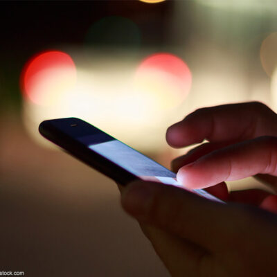 A pair of hands holding a cell phone at night with street lights in the background.