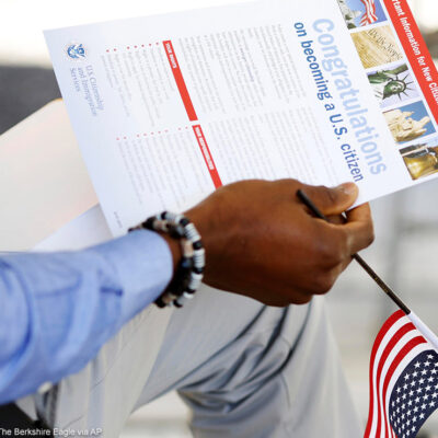 The hand of a seated person holding a miniature U.S. flag and immigration information at a naturalization ceremony.