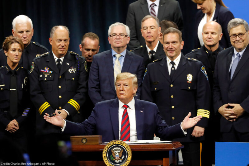 President Trump sits at podium ready to sign executive order creating a commission to study law enforcement and justice, surrounded by officers beside him in support.