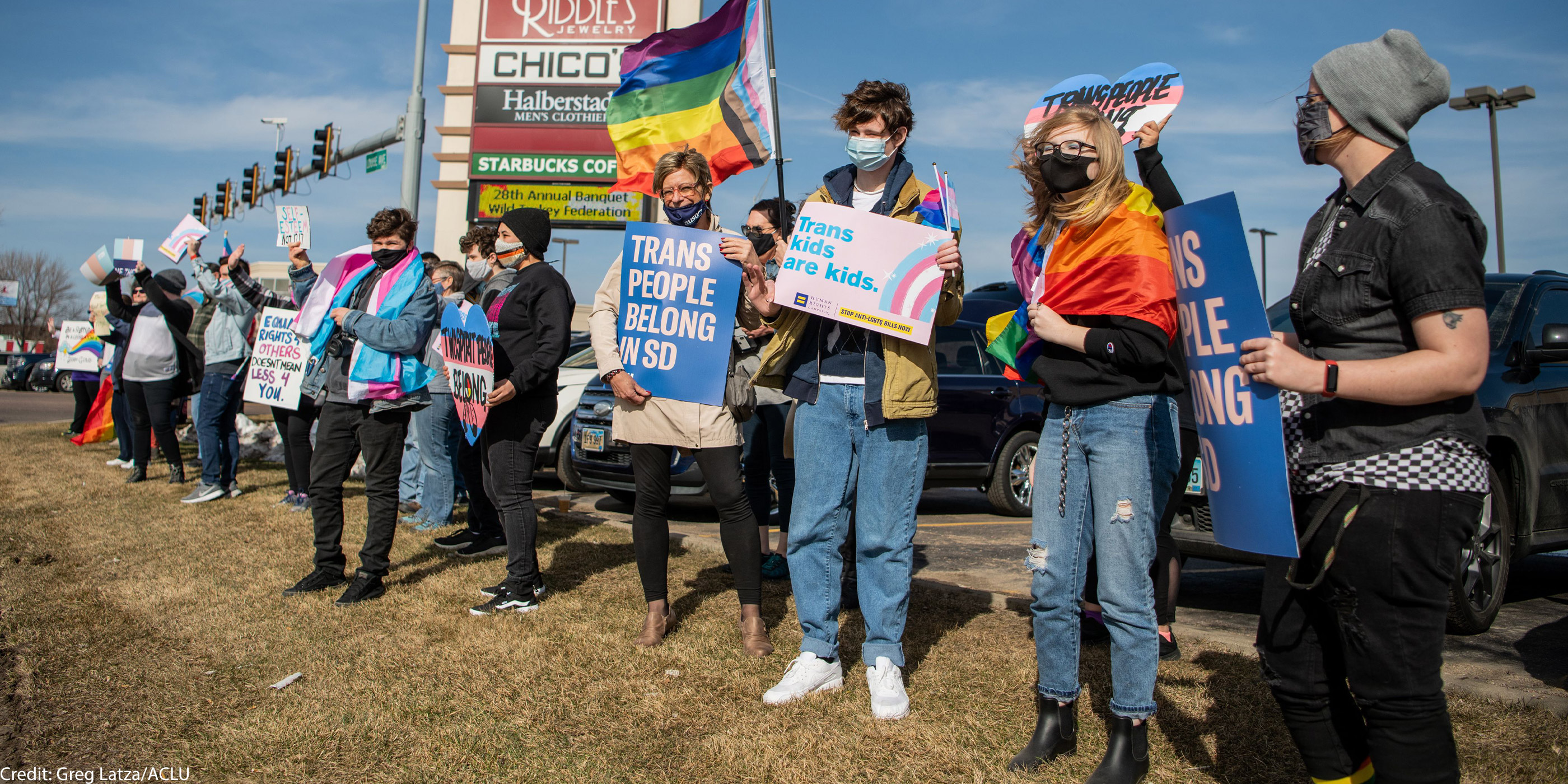Protestors holding an ACLU "Trans People Belong" poster and "trans kids are kids" poster at a trans rights rally in South Dakota.
