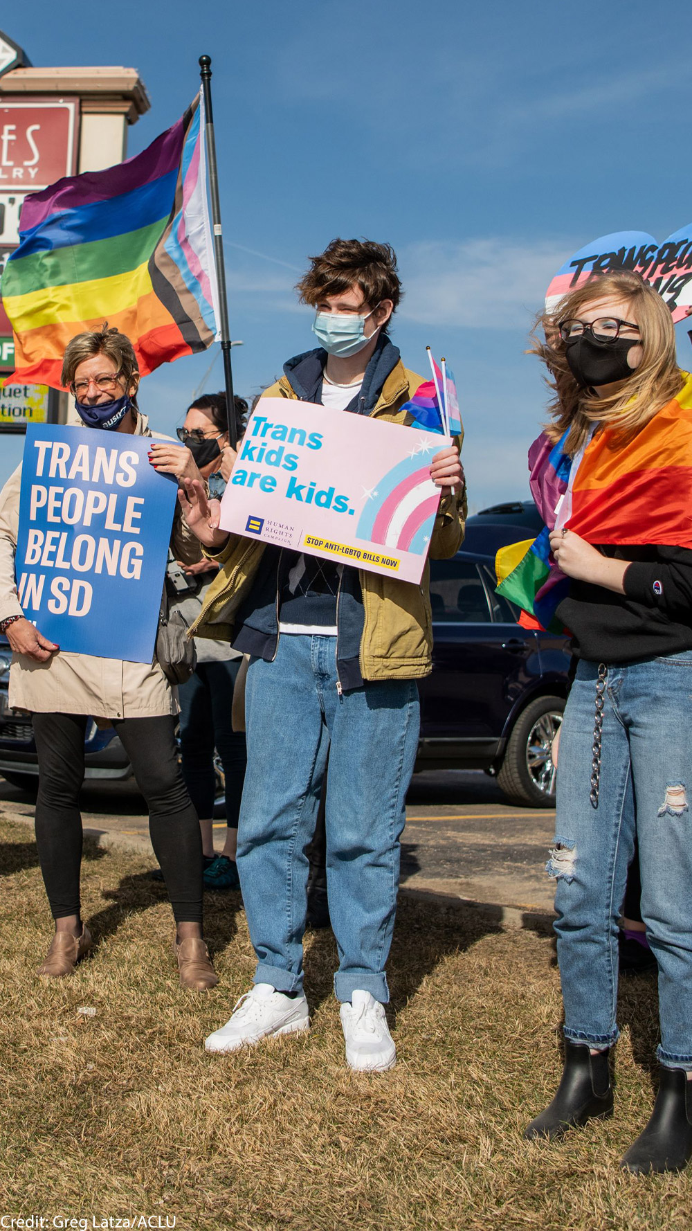 Protestors holding an ACLU "Trans People Belong" poster and "trans kids are kids" poster at a trans rights rally in South Dakota.