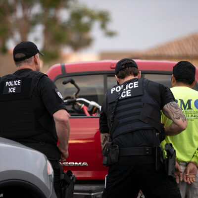 U.S. Immigration and Customs Enforcement (ICE) officers detain someone in front of a red truck.