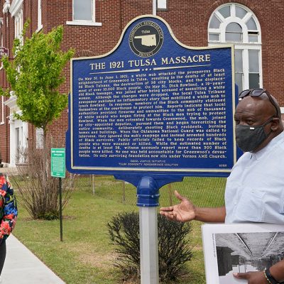 Two people explaining a sign that reads "the 1921 Tulsa Massacre" commemorating the original Black Wall Street
