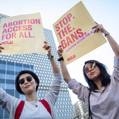 Two women holding up signs that say, "Abortion access for all." and "Stop. The. Bans." respectively.