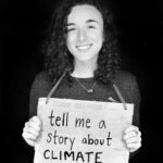 Black and white photo of Devi Lockwood holding a cardboard sign with the words "tell me a story about climate" on it.