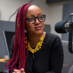 Imani Gandy sits in front of a microphone in a gray cubicle