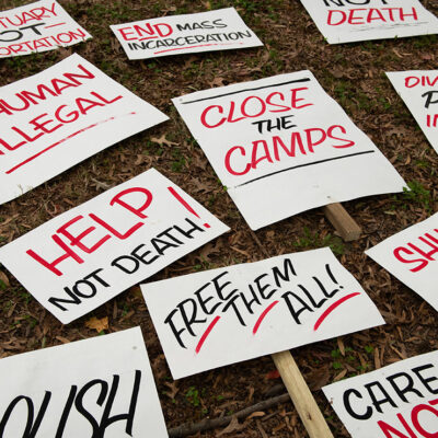 A group of signs on the ground protesting harmful immigration policies, some of which read “No Human Is Illegal,” and “Help Not Death.”