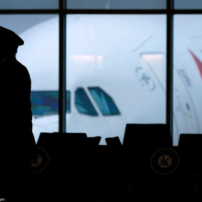 The silhouette of a pilot wearing a facemask walking past a window with a Delta plane outside.