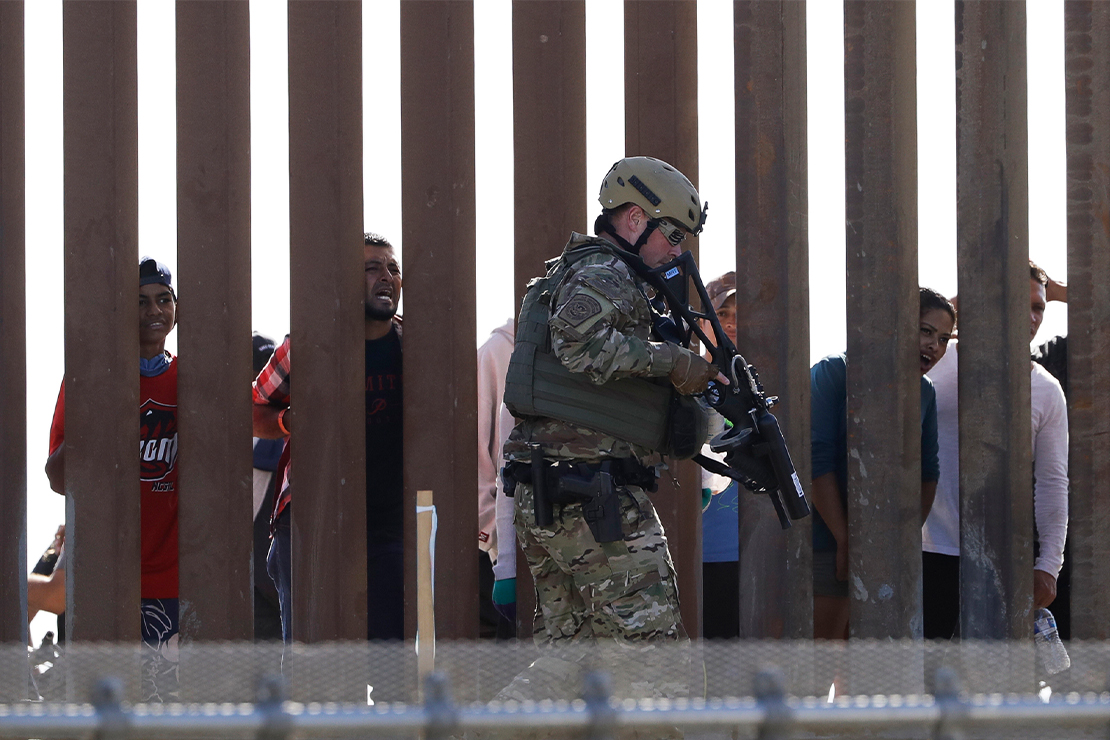 A U.S. Customs and Border Protection officer, with rifle in hand, walks along US side of the border wall as people stare from the opposite side.