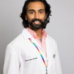 Headshot of man with dark brown/black hair wearing a pink shirt and a white doctor coat. He's also wearing a rainbow colored lanyard.