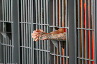 A prisoner holding his extended and clasped hands between the bars of his cell.