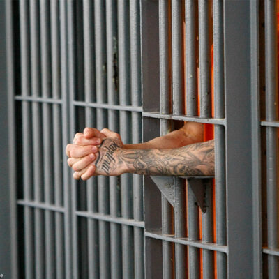 A prisoner holding his extended and clasped hands between the bars of his cell.
