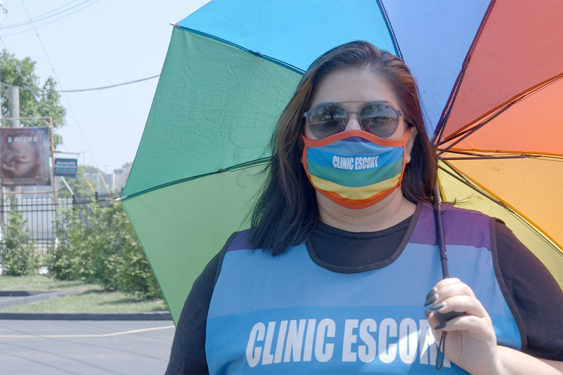 Abortion clinic escort Mariceli Alegria under the shade of an umbrella and wearing a rainbow facemask with the printed words "Clinic Escort".