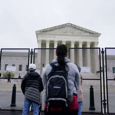 Standing beyond security gates that have signs reading "Area Closed", several people stare at the Supreme Court building.