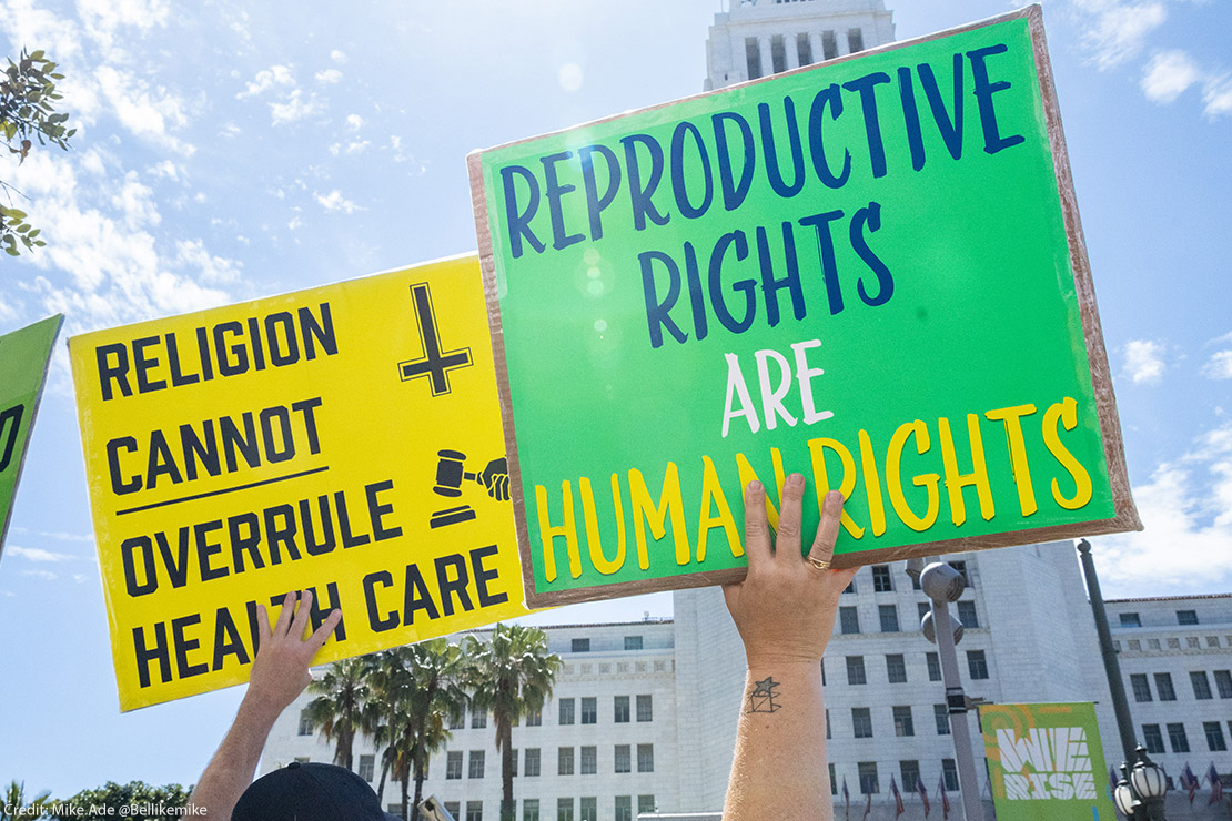 Two protestors at a pro-abortion march holding signs reading "Religion Cannot Overrule Health Care" and "Reproductive Rights Are Human Rights."