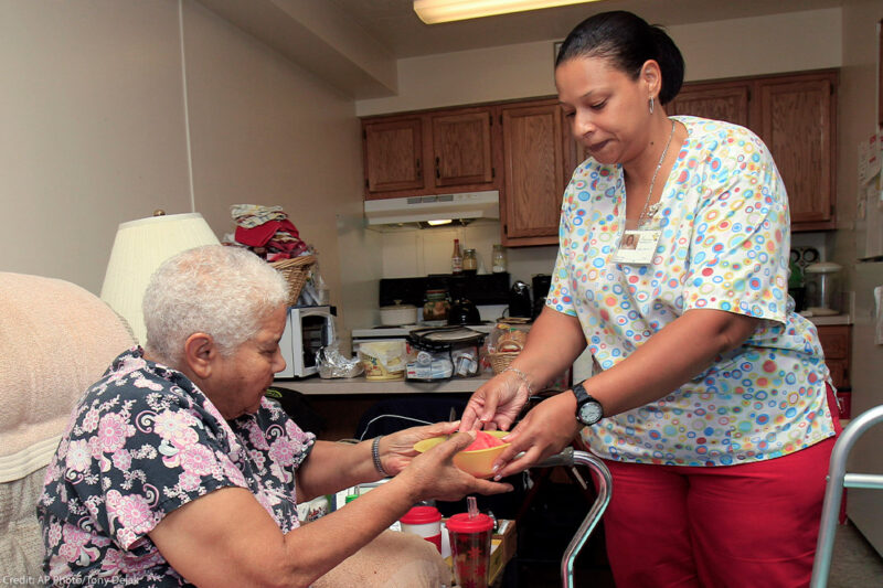 A home health care aide handing her patient a bowl of food.