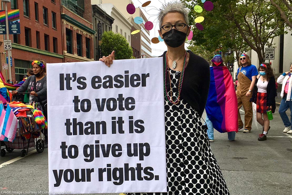 A masked Connie Jeung-Mills in a polka-dotted dress is holding up a sign saying" It's easier to vote than it is to give up your rights.," as people march in the background.