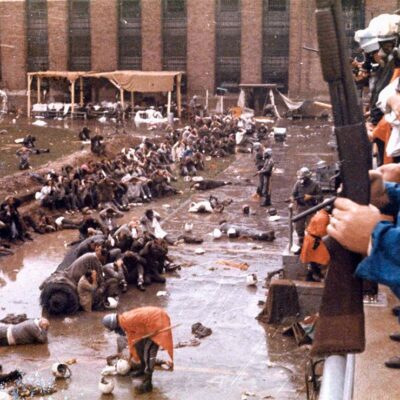 With helmets, gas masks and armed with shotguns, state police overlook the Attica prison yard which is filled with prisoners, face down in mud with their hands behind their heads. Other policemen are in orange ponchos are walking and searching the prisoners in Sept. 1971.