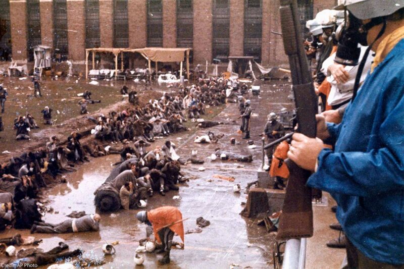 With helmets, gas masks and armed with shotguns, state police overlook the Attica prison yard which is filled with prisoners, face down in mud with their hands behind their heads. Other policemen are in orange ponchos are walking and searching the prisoners in Sept. 1971.