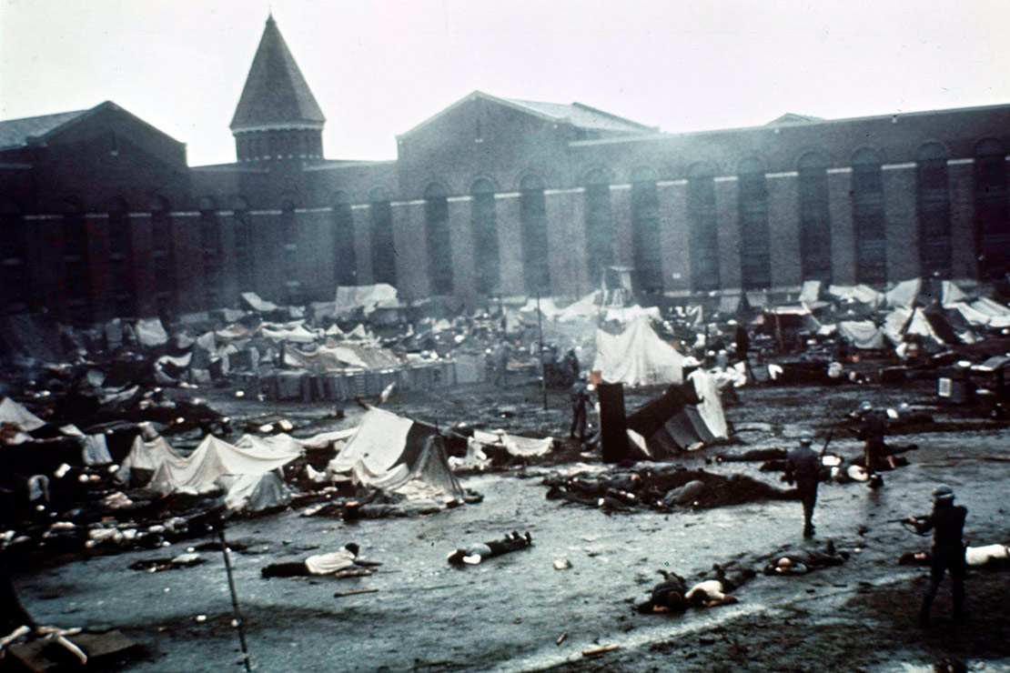 The Attica prison yard is littered with broken make-shift tents, pieces of cardboard and prisoners. The prisoners are forced to lay face down in the mud while state police point towards them with firearms.